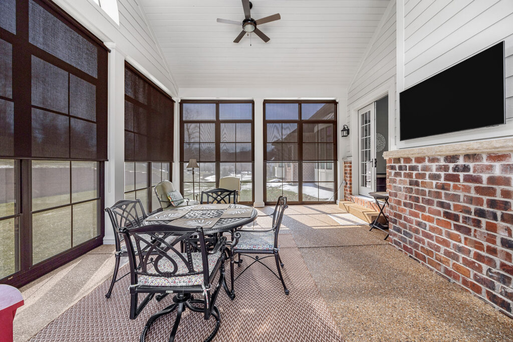 Spacious screened-in porch with retractable solar shades, brick fireplace, wall-mounted TV, ceiling fan, and outdoor dining set.
